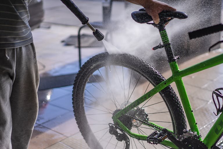 A man cleans a green e-bike with a hose, focusing on the wheel and frame outdoors.